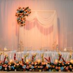 Wedding head table setup with floral arrangements, candles, draped backdrop, and a white frame arch