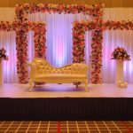 Wedding stage with floral arch frames, golden tufted sofa, pedestal flower vases, and warm candlelight