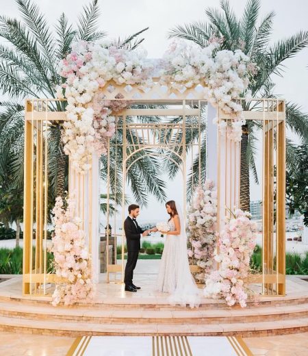 Bride and groom exchanging vows under a floral wedding arch with gold frame in an outdoor ceremony