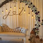 Wedding stage with cream sofa, floral arch, hanging lights, and personalized initials backdrop
